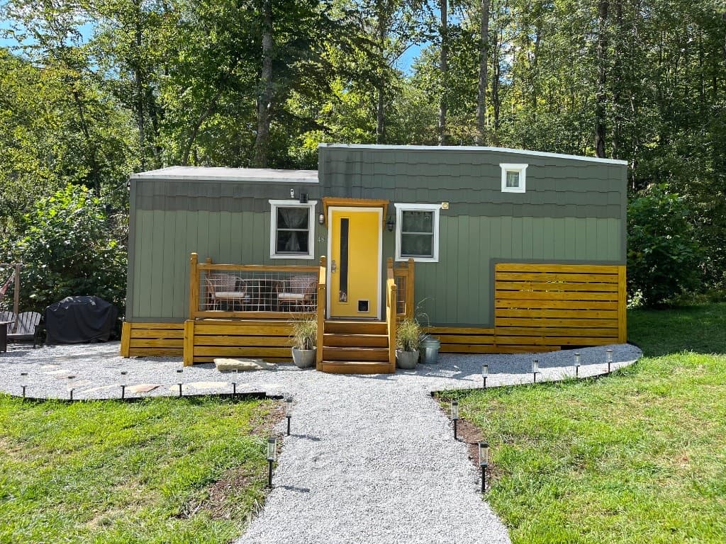 Olive-green tiny cabin with a yellow front door, wooden deck, and gravel path in the woods