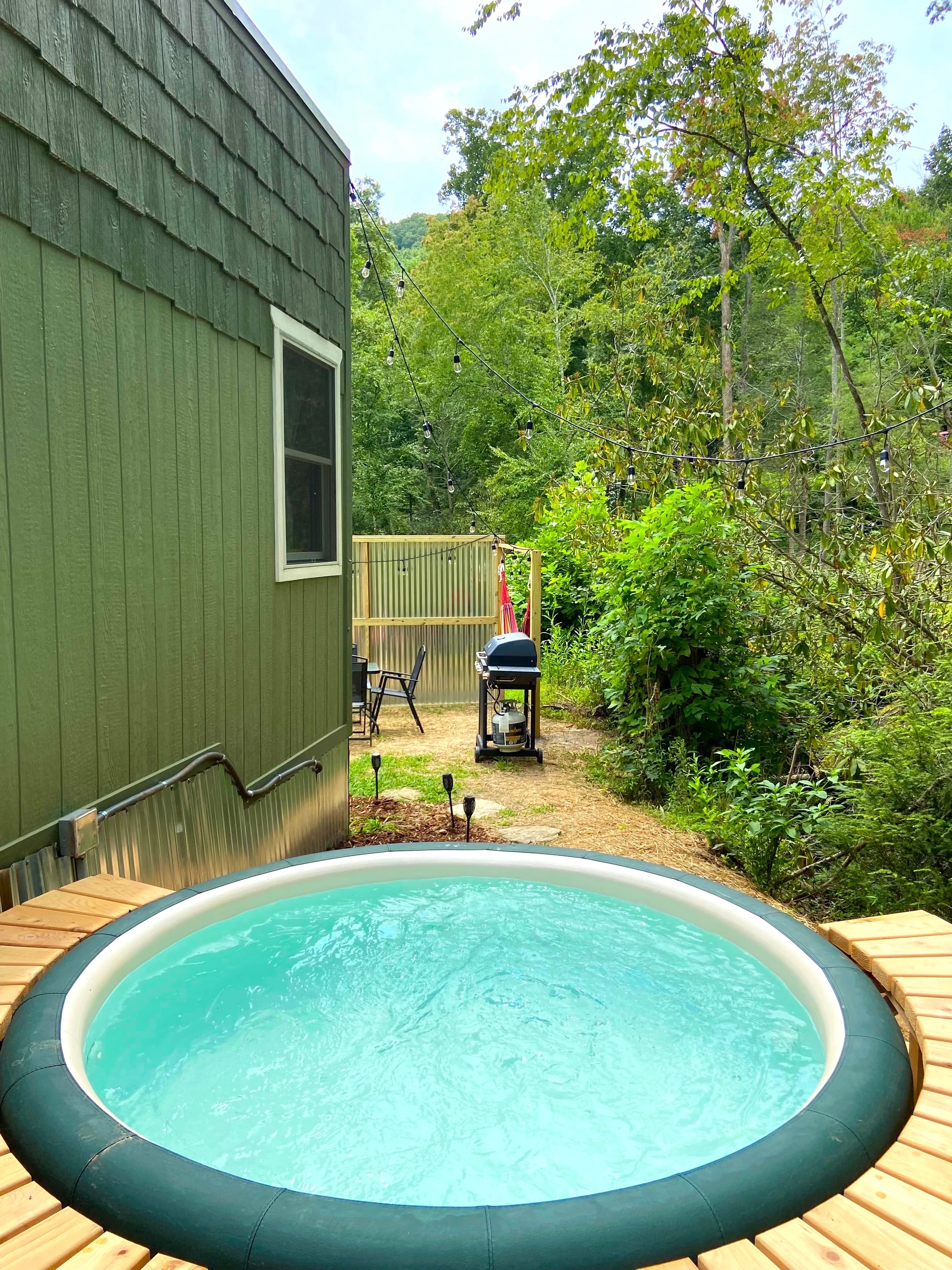 Open private hot tub on a wood deck with grill, dining, and string lights through the trees